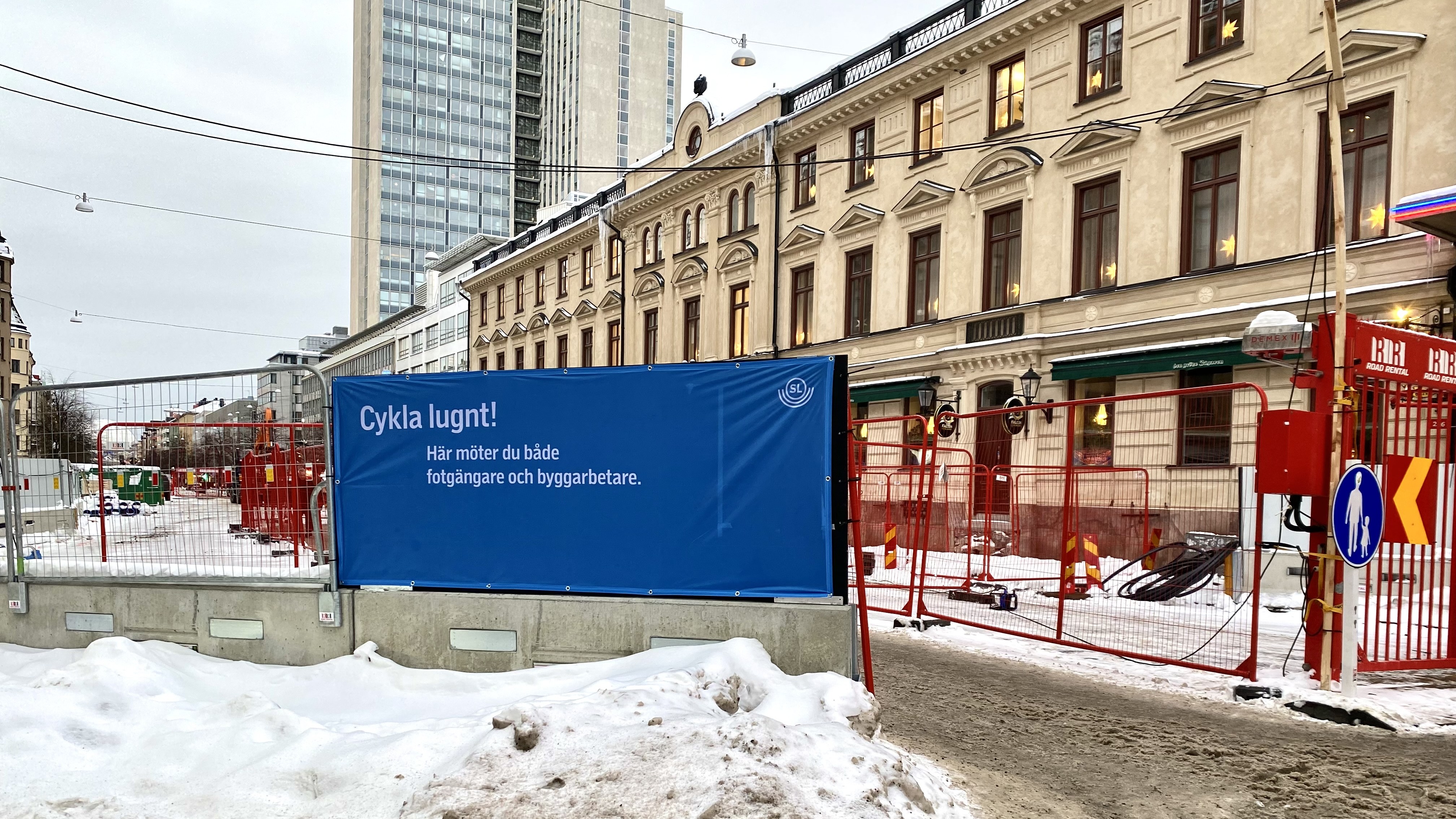 En snöig gata med avspärrning för byggarbetsplats med gallerstängsel. På en blå stor skylt står: Man har satt upp en stor blå skylt på en av byggarbetsplatsens gallerstaket där det står: Cykla lugnt, här möter du både fotgängare och byggarbetare