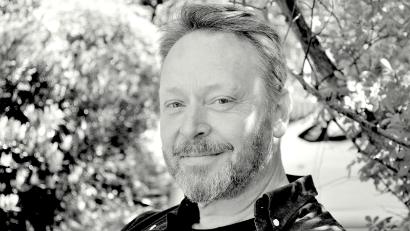 Svartvitt foto. Vit medelåldersman med kort hår och kort skägg, sitter med grönskande buskar runt sig.