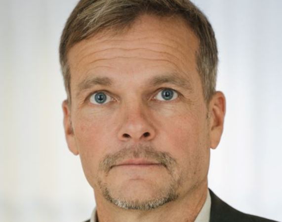 Vit medelålders man med kårt gråblont hår med snedbena. Blå ögon, grå mustasch. Vit skjorta, grå kavaj.