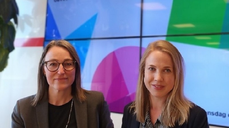 Två kvinnor framför en skärm med MFD:s logotyp. En är medelålders med mörkbrunt axellångt hår och glasögon, och en är något yngre med blond halvlång frisyr.