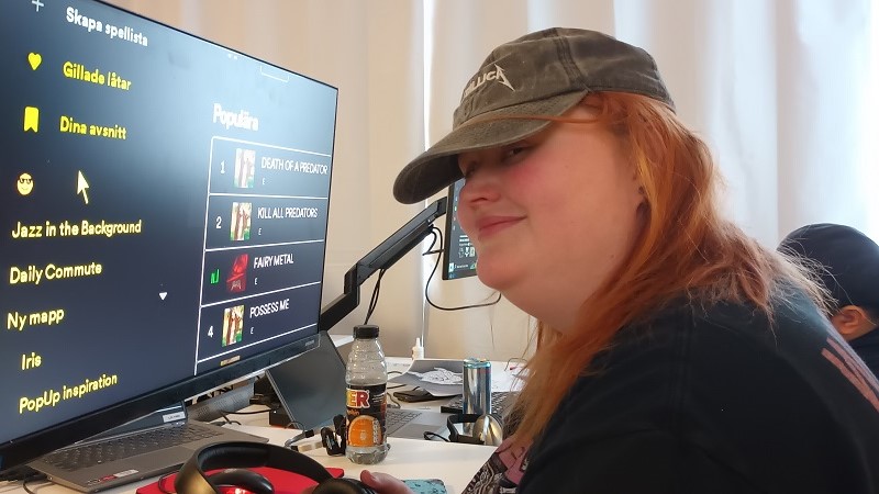 Ljus ung kvinna med keps, mellanlångt rött hår. Sitter vid stor datorskärm.