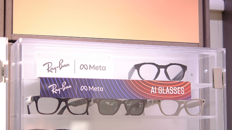 På en butikshylla ligger glasögon, de har små runda kameralinser i skalmarna. Bredvid glasögonen finns skyltar med logotyper för Ray-Ban och Meta.