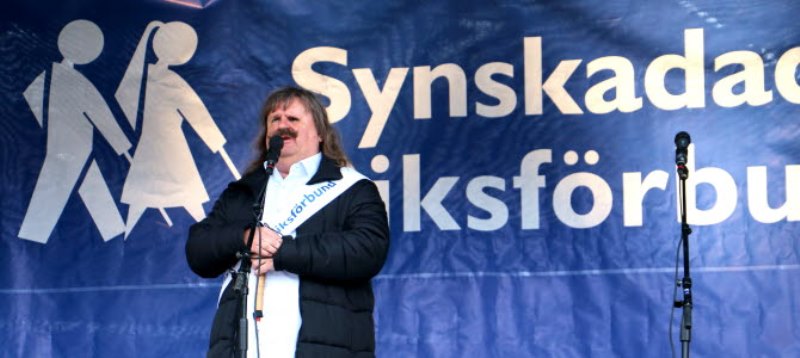 Gråhårig man står framför en stor SRF flagga i blått med vit text