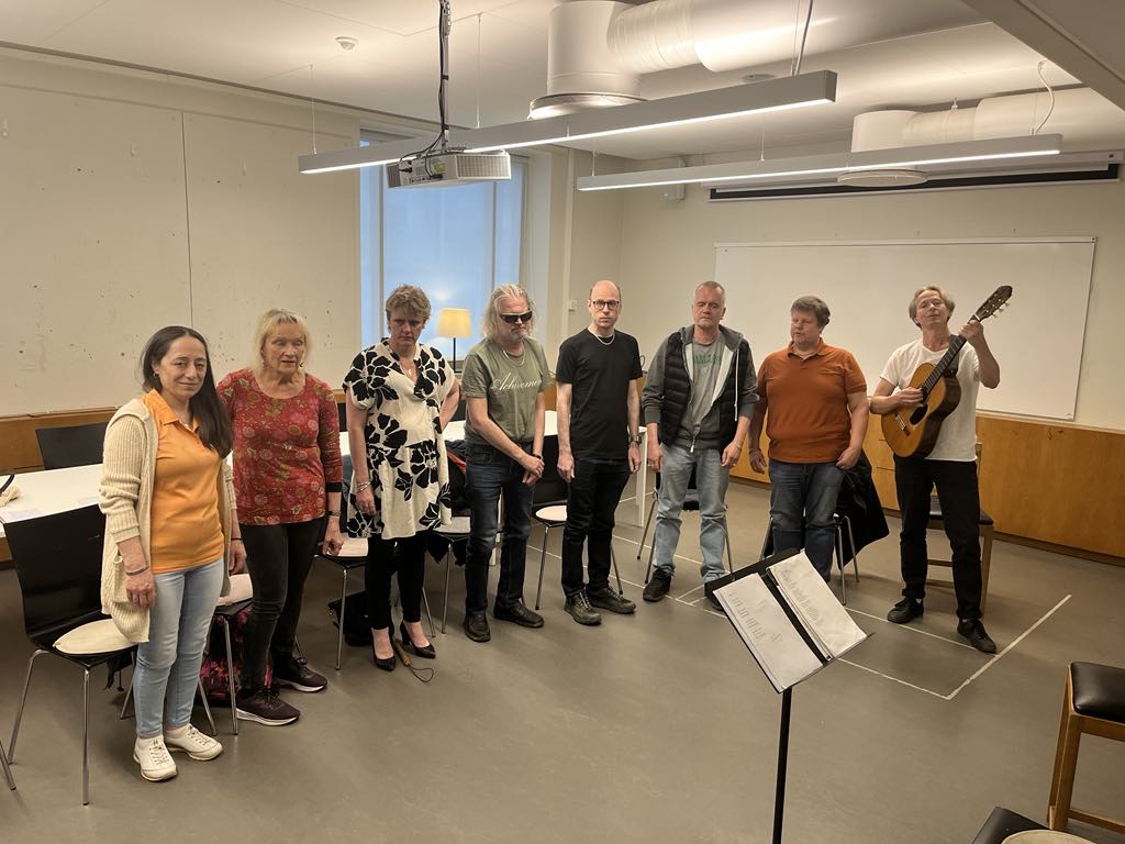 I en replokal står en grupp medelålders personer bredvid varandra, en av dem har en gitarr.