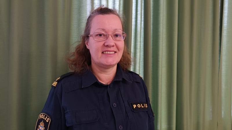 En vit medelålders kvinna med halvlångt lockigt brunt hår, glasögon och polisuniform.