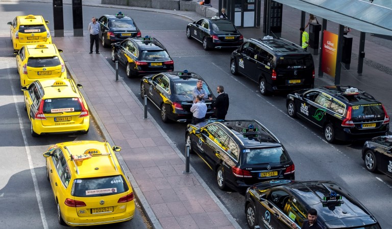 Tre rader parkerade taxibilar vid en hållplats. Gula bilar på rad i vänsterkant, resten är svarta. Några förare står utanför bilarna och väntar.