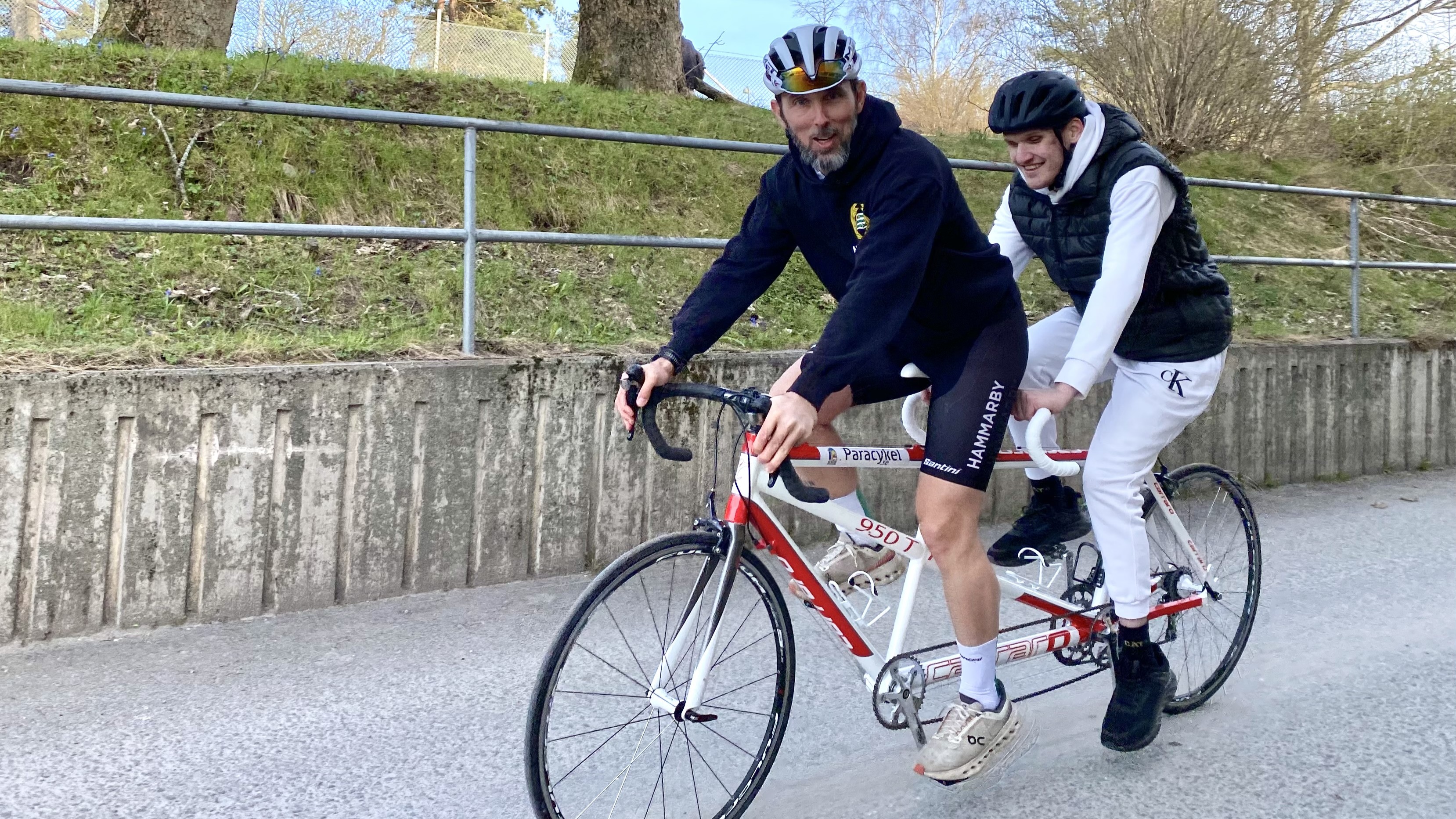 Två män cyklar på en vit tandemcykel i parkområde. Den ena har blå jacka och korta cykelbyxor, den andra vit träningsoverall. 