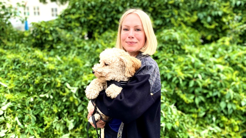 Ljus, ung blond kvinna med page med beige pudel i famn, står i grön park.