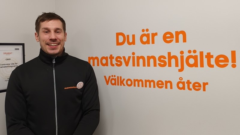 En vit man med brunt lätt rufsigt hår klädd i svart står vid en vägg med texten ”Du är en matsvinnshjälte! Välkommen åter”