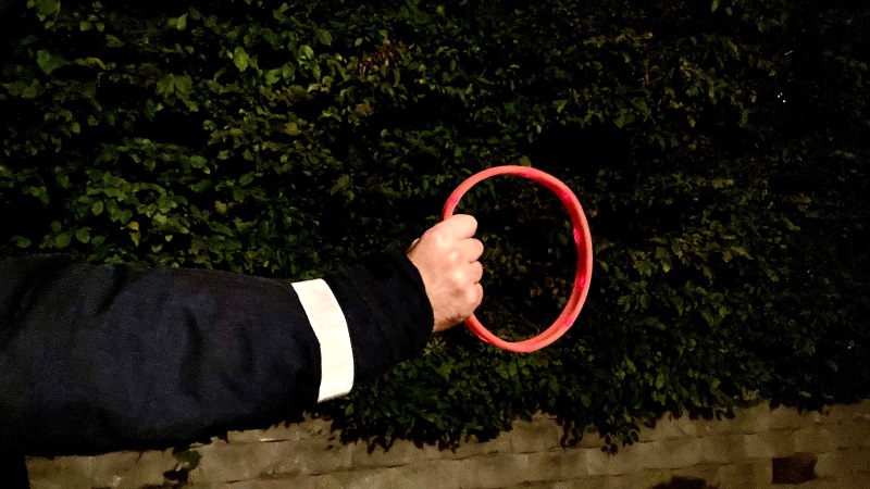 Närbild i mörk utemiljö, utsträckt arm i mörk jacka med vit reflexrem, i handen hålls ett rött reflexhalsband för hund.