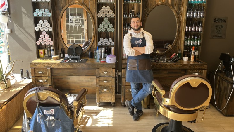 En 30-årig man från mellanöstern, med mörkt kort hår och kort skägg. Har vit hoodie, jeans, läder förkläde. Står i barberarsalong med bruna trähyllor.  