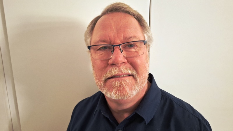 Medelålders vit man med grått hår och skägg. Har mörkblå skjorta. Vit bakgrund.