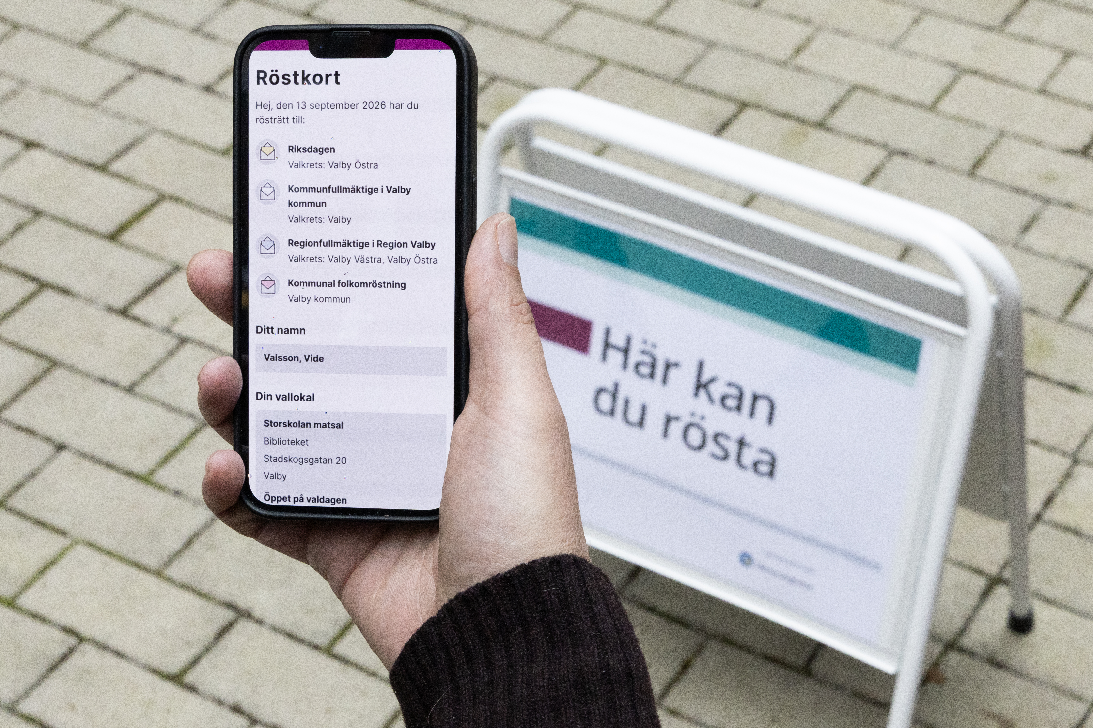 I flrgrunden en hand som håller en smart telefon med ett digitalt röstkort som syns på skärmen där man kan läsa text om namn och vallokal. I bagrunden en gatupratare med texten "här kan du rösta"