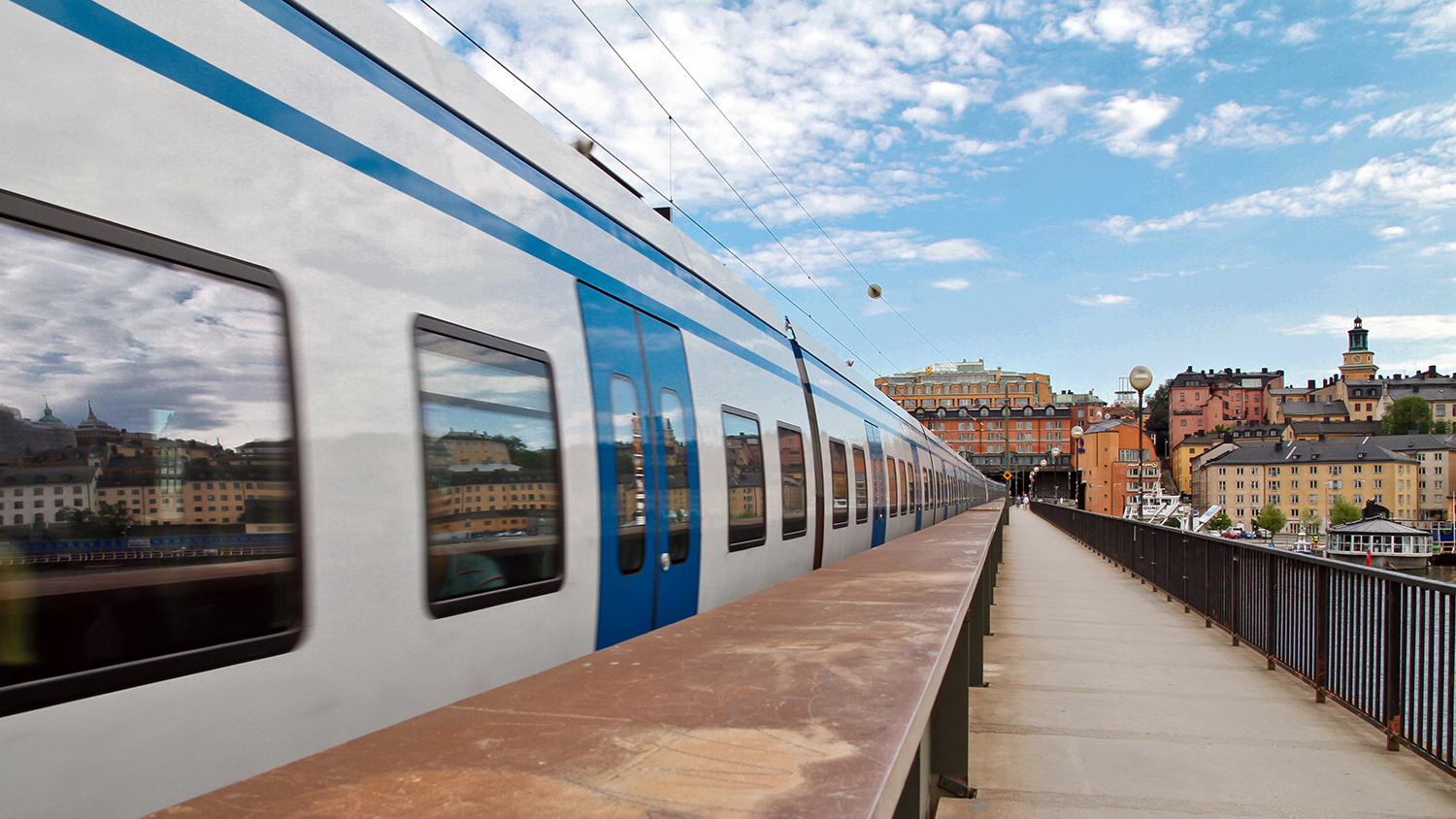 Ett åkande ljusgrått pendeltåg på bron mot Gamla stan. Blå himmel.