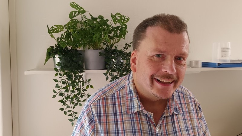 En leende vit medelålders man med ljusbrunt hår, gråblå ögon och mustasch.
