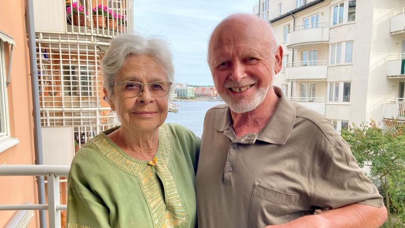Ett äldre gråhårigt par med ljus hy, håller om varandra på balkongen. Vy med hyreshus och sjöglimt.