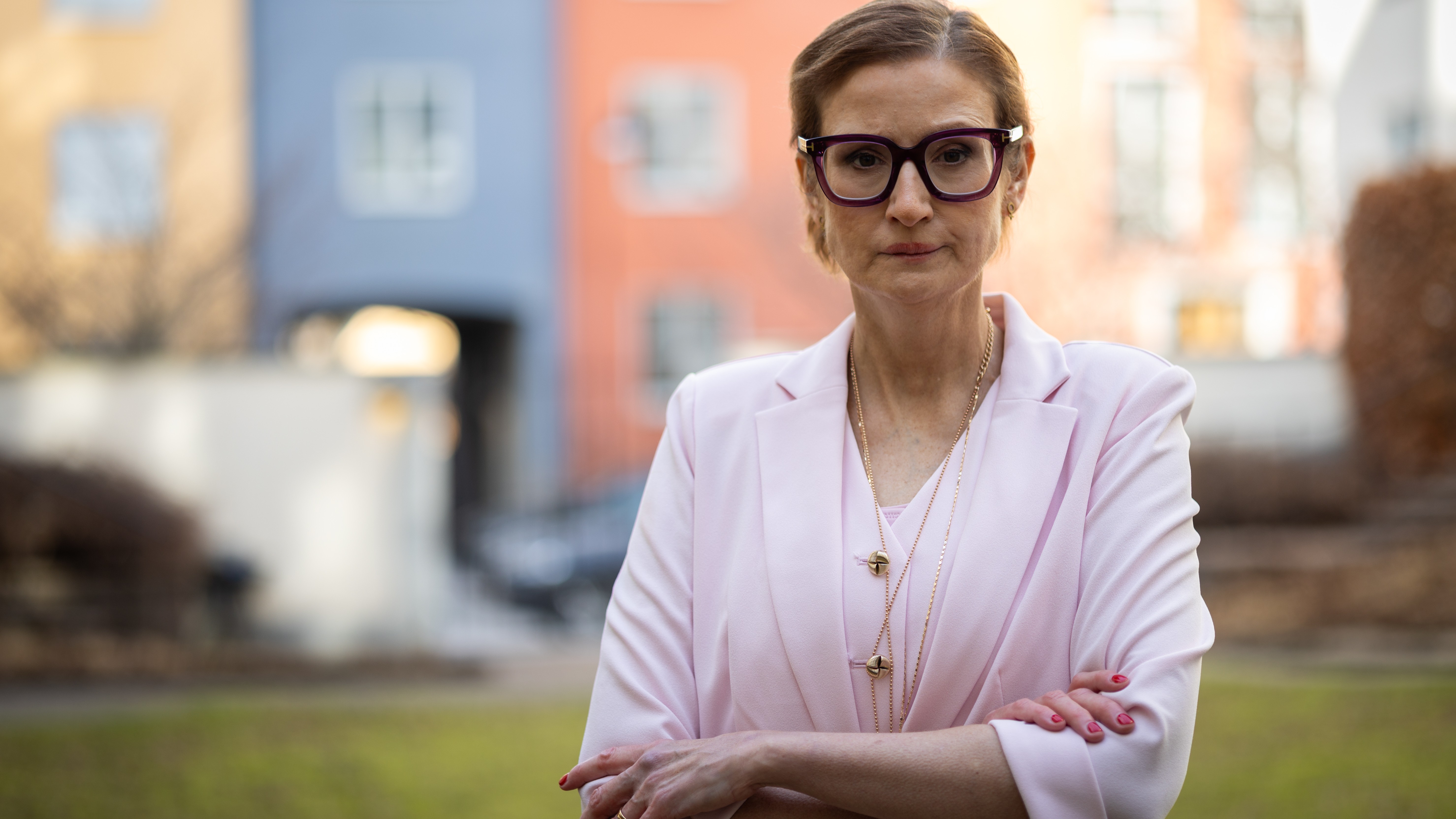 Vit slank medelålders vit kvinna med kort brunt hår, stora mörkbågade glasögon. Ljusrosa kostym. Står framför några gatuhus i gult, rostrött och blått.