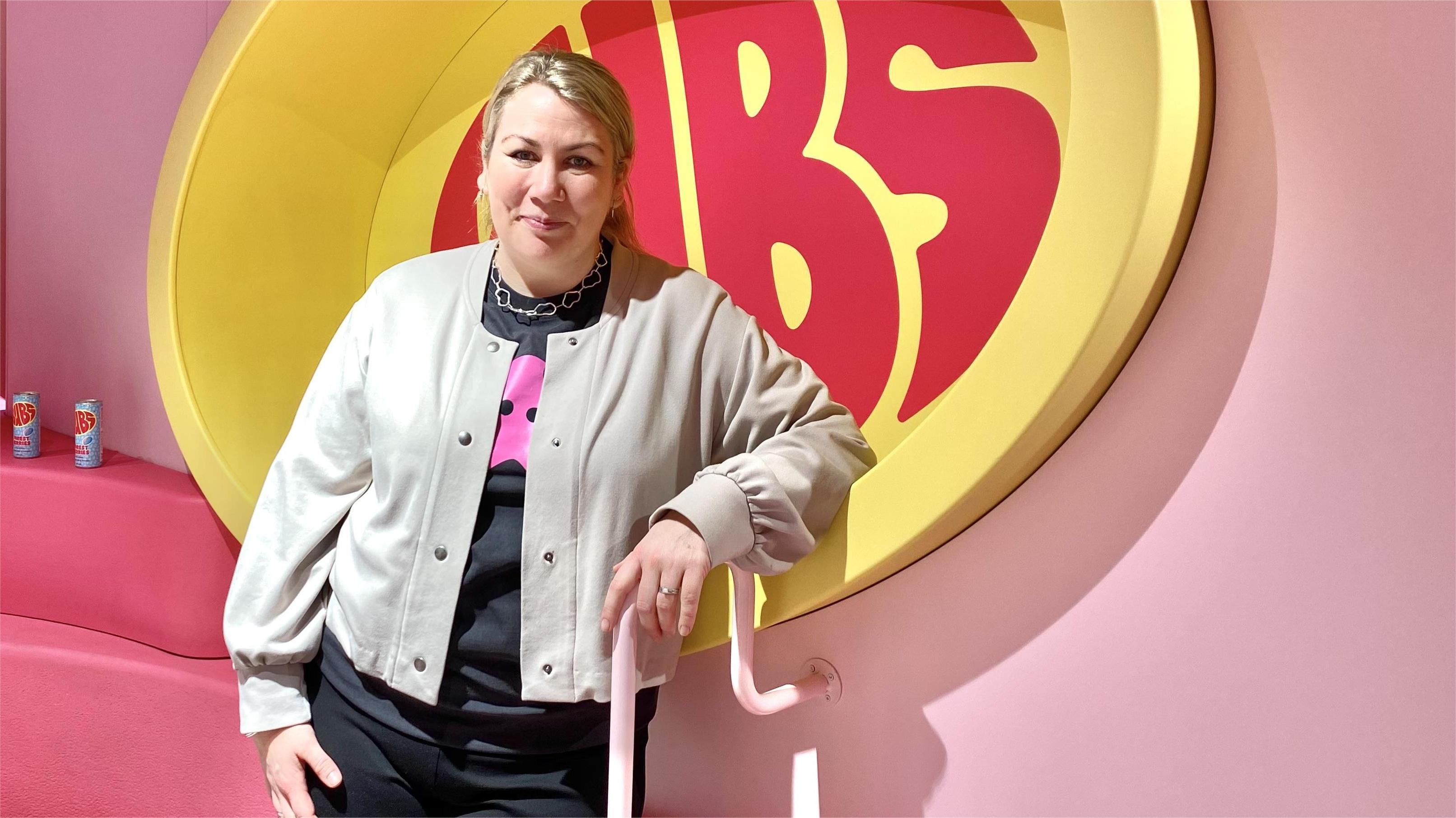 Joanna Mossberg, kommunikationschef BUBS. 35 årig kvinna med blont hår i tofs, beige kofta och svart t-shirt under. Står vid rosa vägg med stor logotype av ”BUBS”- rosa bokstäver i ljusgul oval.