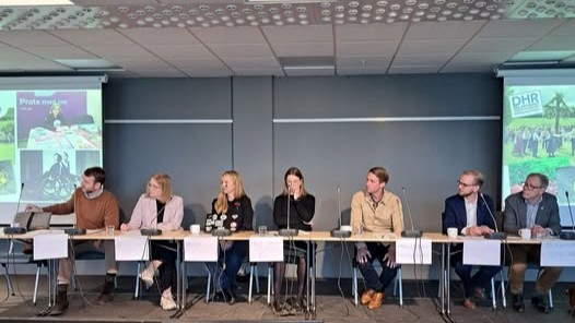 Längs ett avlångt bord sitter flera politiker på rad bredvid varandra. Stor samlingssal med två stora tv-skärmar i bakgrunden.