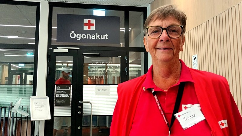 Framför en ingång märkt ”Ögonakut” står en kortklippt brunhårig kvinna med glasögon och ljus hy i röda kläder och en namnskylt det står Yvonne på. 