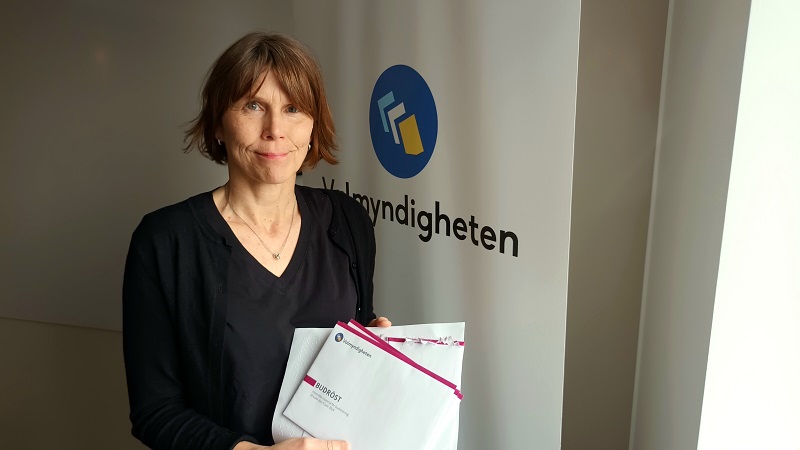 Lotta Liedberg, en smalt byggd vit medelålders kvinna med kastanjebrunt hår i page och med rak näsa, håller i kuvert med texten budröst, framför en banderoll med Valmyndighetens logga.s