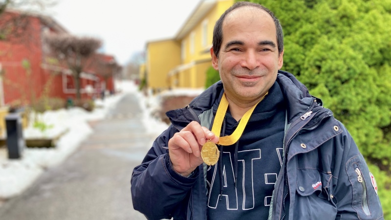Medelålders man med svart snaggat hår, svart jacka, håller fram en guldmedalj han har runt halsen. Radhusgata bakom.