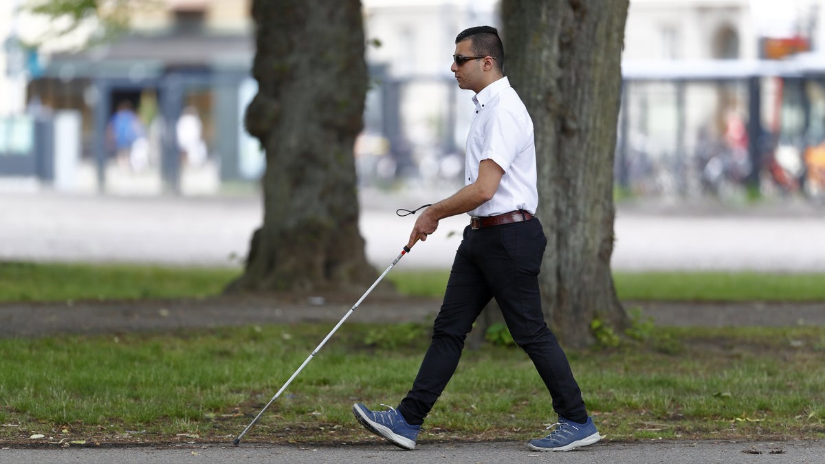 En yngre man med kort mörkt hår och brun hy går med sin vita käpp på trottoar. Han har vit t-tröja och svarta jeans. Vägen ligger vid grönområde med gröna träd. Sommar.