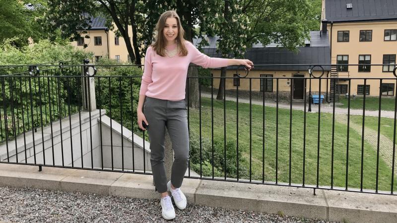 Kvinna med halvlångt brunt hår står vid staket i en grönskande park.