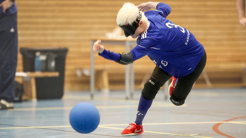En person med blont hår, svart ögonbindel och blå idrottströja har just kastat iväg en blå goalball.
