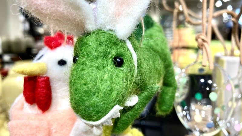 En liten grön tygdinosaurier hänger i ett påskris, även en vit tyghöna och et genomskinligt glasägg
