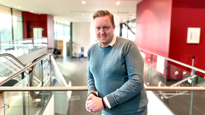 Vit medelålders man med gråblå långärmad tröja, står i väldigt stor entré med rulltrappor och stora fönster