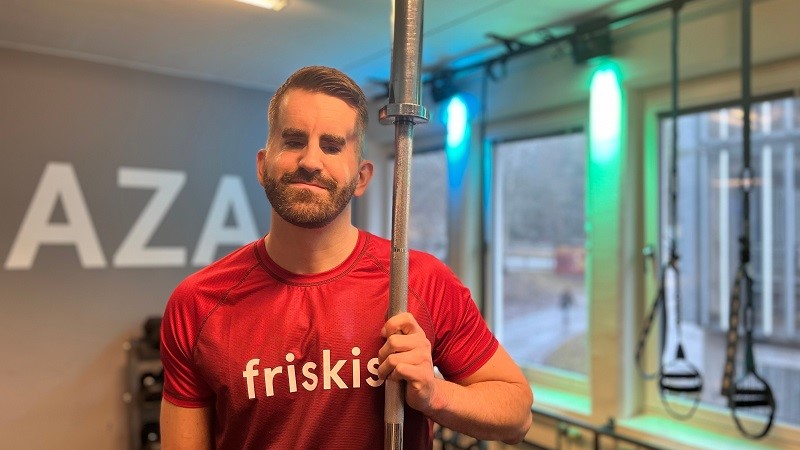 En ljushyad muskulös man med brunt skägg, i röd t-shirt med texten FIRSKIS i vitt, håller i en upprätt skivstång.