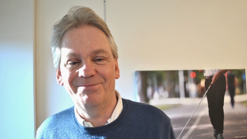 Medelålders leende man i närbild, kort grått hår, mörkblå tröja, vit skjortkrage syns
