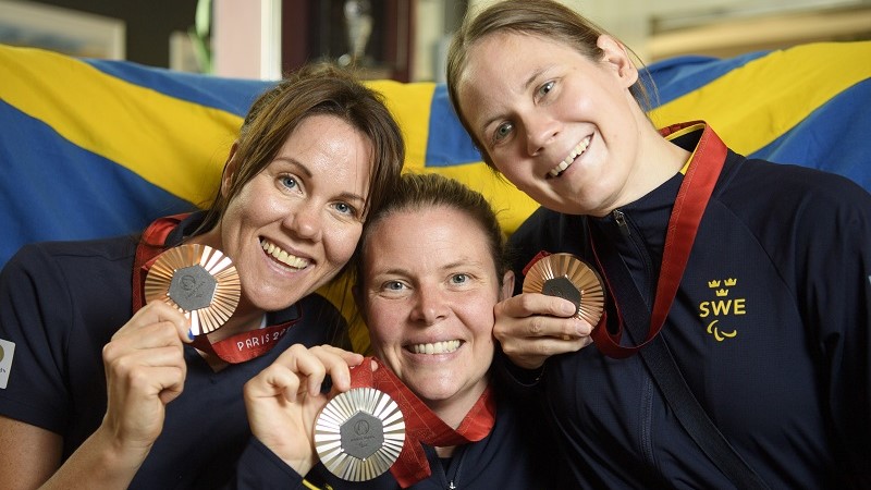 Tre vita kvinnor ler och håller upp varsin medalj, en svensk flagga i bakgrunden.