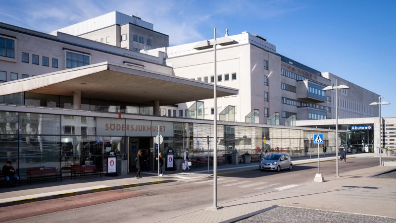 Södersjukhusets huvudentré . En låg byggnad med fönster längs hela fasaden, framför går en väg med ett övergångsställe. Ovanför entrén står det SÖDERSJUKHUSET i en orange nyans. I Bakgrunden flera grå sjukhusbyggnader i varierande höjd.