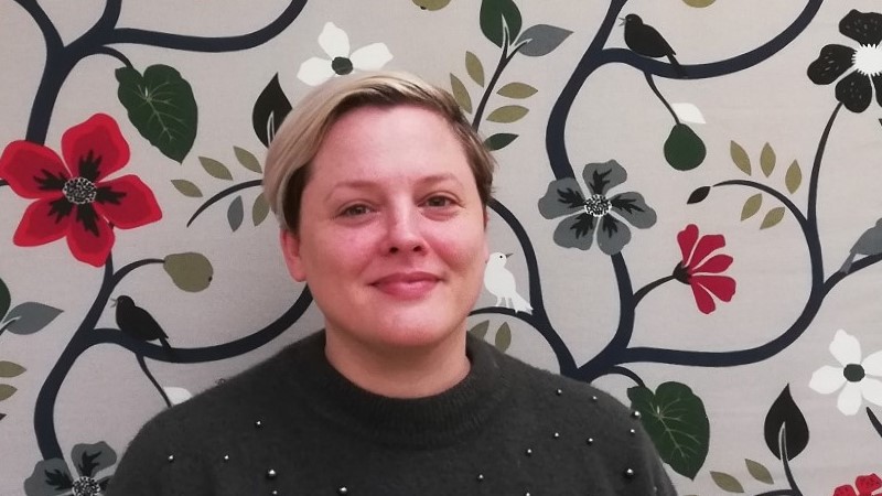 En kvinna med ljus hy och blont hår med mörka rötter står framför en mönstrad bakgrund med blommor och fåglar.
