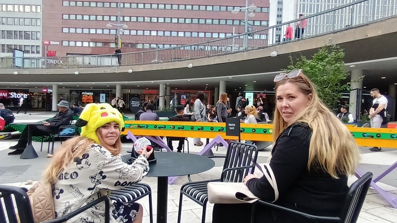 Ett barn med en gul pikachu-mössa och hennes mamma sitter på varsin stol på sommartorget. Runt om finns spel, andra sittplatser och människor.