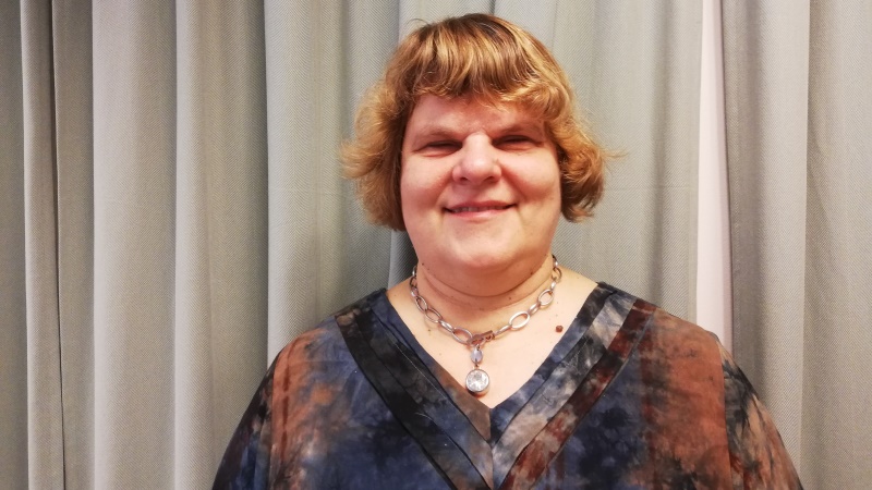 Medelålders rödlätt ljus kvinna, kort hår, brunmelerad blus, framför grått draperi.