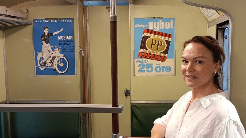En ljushyad kvinna i mörkblont hår och vit blus står i en gammal tunnelbanevagn med reklamskyltar för Mustangmoped och PP Lakritstabletter.