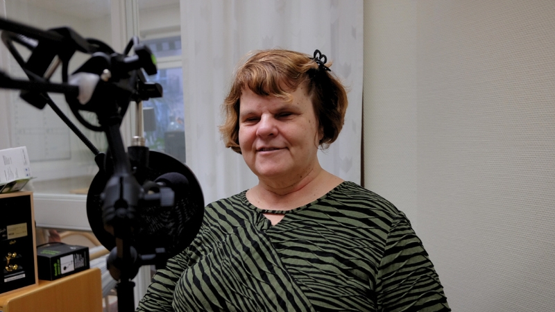 Karin Hjalmarsson, ordförande SRF Stockholm Gotland, en vit kvinna i 50-årsåldern med kort ljusbrunt hår sitter framför en mikrofon. Hon bär en grön- och svartmönstrad tröja och har en liten hårklämma i håret.