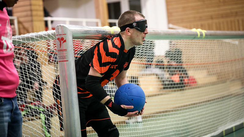 En kille i svart träningsoverall, står i ett goalballmål, håller blå boll i händerna.