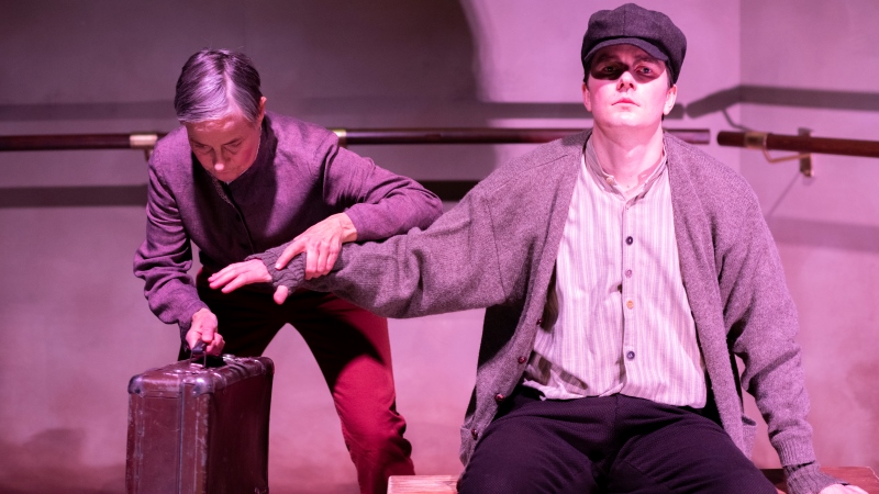På en teaterscen sitter en ung blind man med armen utsträckt där en kvinna tar emot den och håller själv i en resväska.