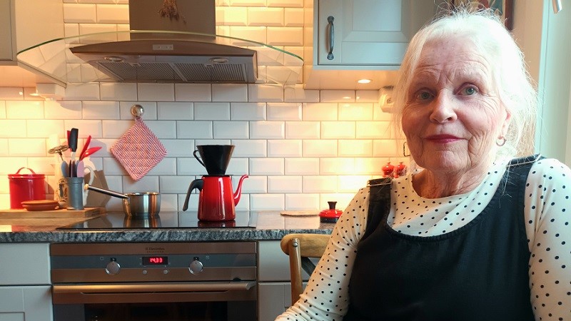 En ljushyad vithårig kvinna med rött läppstift sitter vid ett köksbord. En burk kakor och en kaffekopp står på bordet.