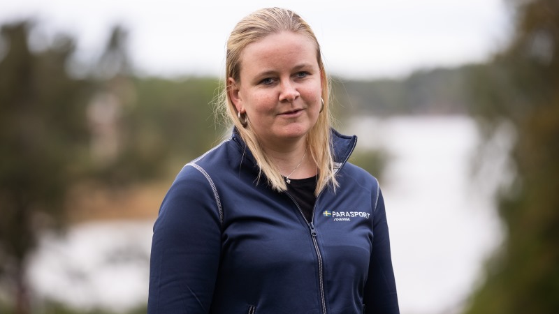 Ung blond ljus kvinna med tofs, i blå träningsjacka står framför skog med snö.