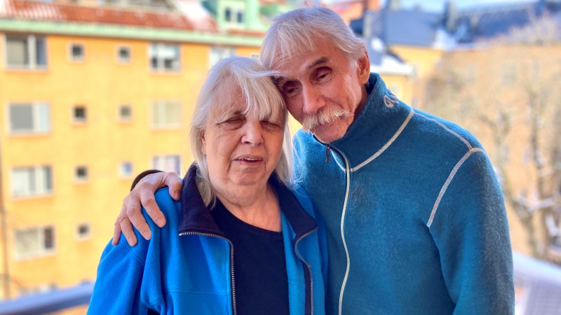 Ett äldre ljushyat par står och håller om varandra på sin balkong med utsikt mot gården och blå himmel. De har turkosa fleecejackor på sig