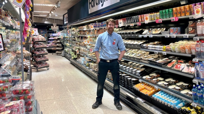 Man i blå skjorta i butik, står mellan två höga matvaruhyllor