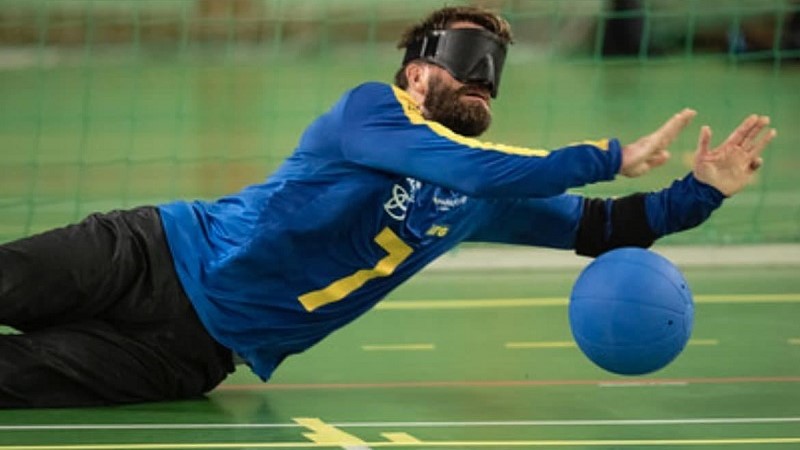 En vit man med skägg och svart ögonbindel, iförd blå landslagströja, sträcker ut sig på marken mot en blå boll.