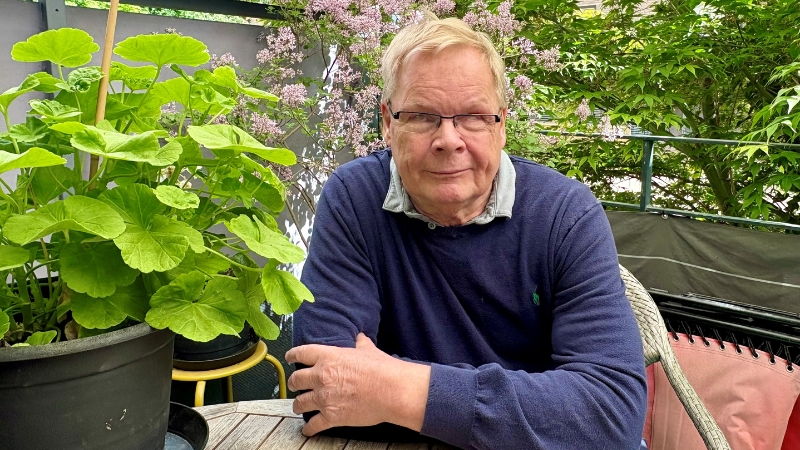vit 70-årig man med kort vitt hår och mörkblå tröja sitter vid ett träbord på sin växtbebodda terrass.