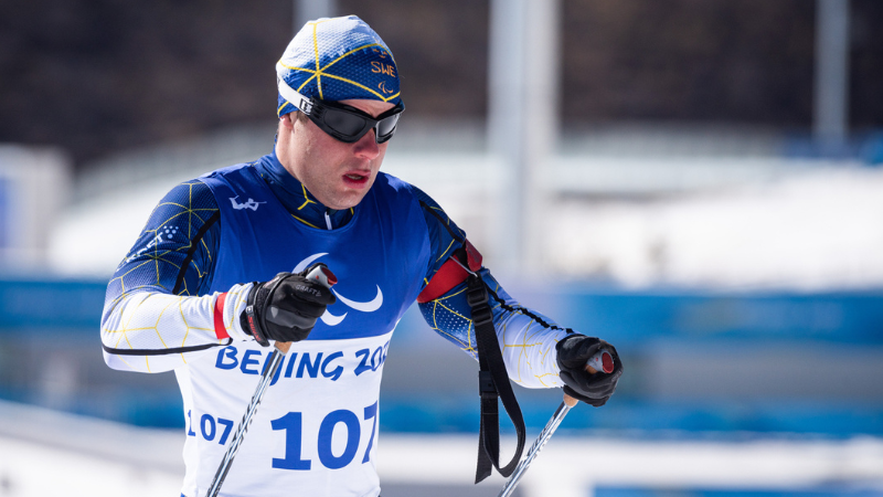 Längdskidåkaren Zebastian Modin i blå mössa, svarta skidglasögon och blåvit dräkt med ett linne på där det står Bejing. I händerna har han stavar. Han är vit man i 30-årsåldern och på bilden är ansiktet koncentrerat.