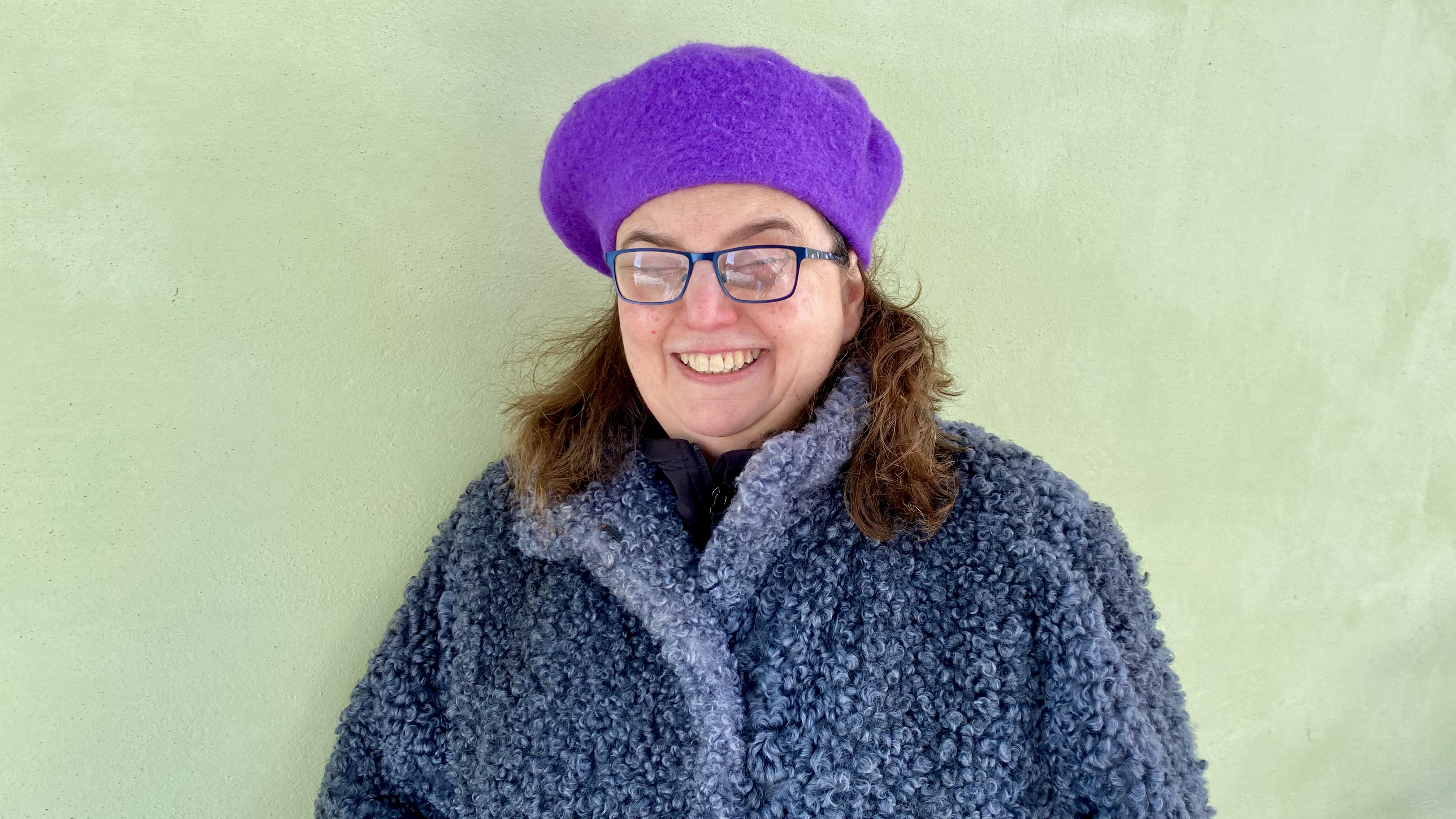 En medelålders vit kvinna med brunt axellångt hår, lila basker och en grå kappa med syntetisk småkrullig fårullspäls. Hon står vid en lindblomsgrön vägg utomhus.